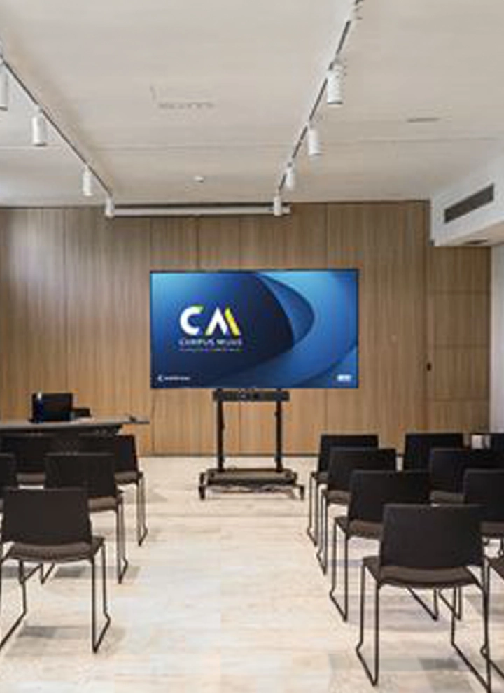 En la foto se ve un monitor que enseña en su pantalla un fondo de diferentes tonos azules sobre el que pone CM, las iniciales de Campus Mijas, la propuesta formativa de ILUNION Hotels. Ante la pantalla se encuentra un pasillo de suelo divisorio de dos filas diferentes de sillas, una a la derecha y otra a la izquierda, ambas compuestas de varias sillas. Las sillas en cuestión son negras con patas de metal.