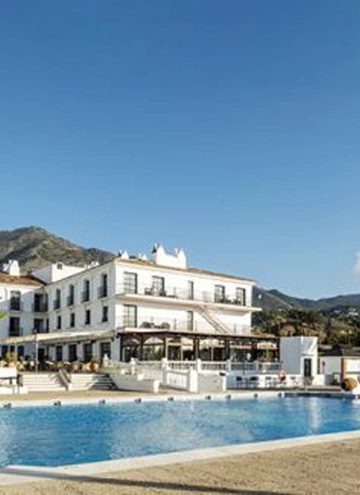 En la fotografía se observa la fachada del hotel ILUNION Hacienda de Mijas. Dicha fachada es blanca al completo, exceptuando los techos que son de un tono marrón oscuro. Frente al hotel se puede ver la piscina que pertenece a este mismo, una piscina grande de agua limpia y azul cristalina. Tras el hotel se puede ver a lo lejos, casi al horizonte, la silueta de un par de montañas. También se puede ver un cielo azul despejado de nubes y Sol.