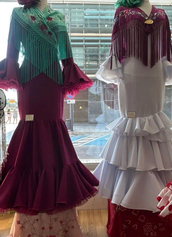 En la imagen se pueden ver dos trajes de flamenca frente a una cristalera. El traje de la izquierda es granate con vuelo en el vestido sobre la mitad de la rodilla, acompañado de un mantón verde agua decorado con flores y una flor del mismo color que el vestido. El traje de la derecha es blanco con un mantón rojo del color del vestido de a izquierda y una flor del verde del mantón del traje anterior.