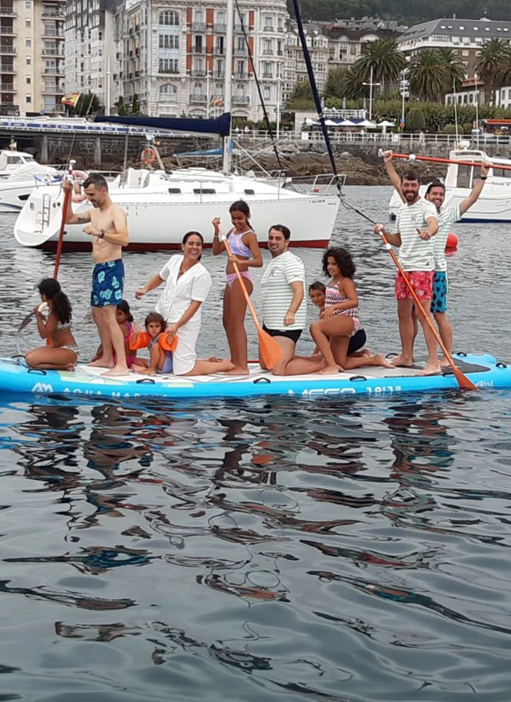 En la foto se ve a un grupo de personas sobre una lancha, algunos visten bikinis y bañadores coloridos mientras otros visten camisetas y pantalones de diferentes tipos. Se puede ver el mar sobre el que están navegando, al fondo de la imagen se observa un par de barcos pequeños blancos mientras que sobre la lancha que ellos están es naranja con bordes azul claro. Todos posan mirando a la cámara sonrientes, incluso algunos cogen los remos.