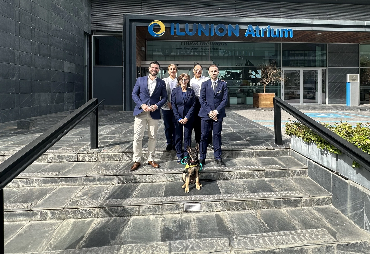 EL equipo de ILUNION Atrium, posa junto a Lia, primera perra guía criada por un hotel. Posan frente a la entrada de ILUNION Atrium con una sonrisa en un día soleado.