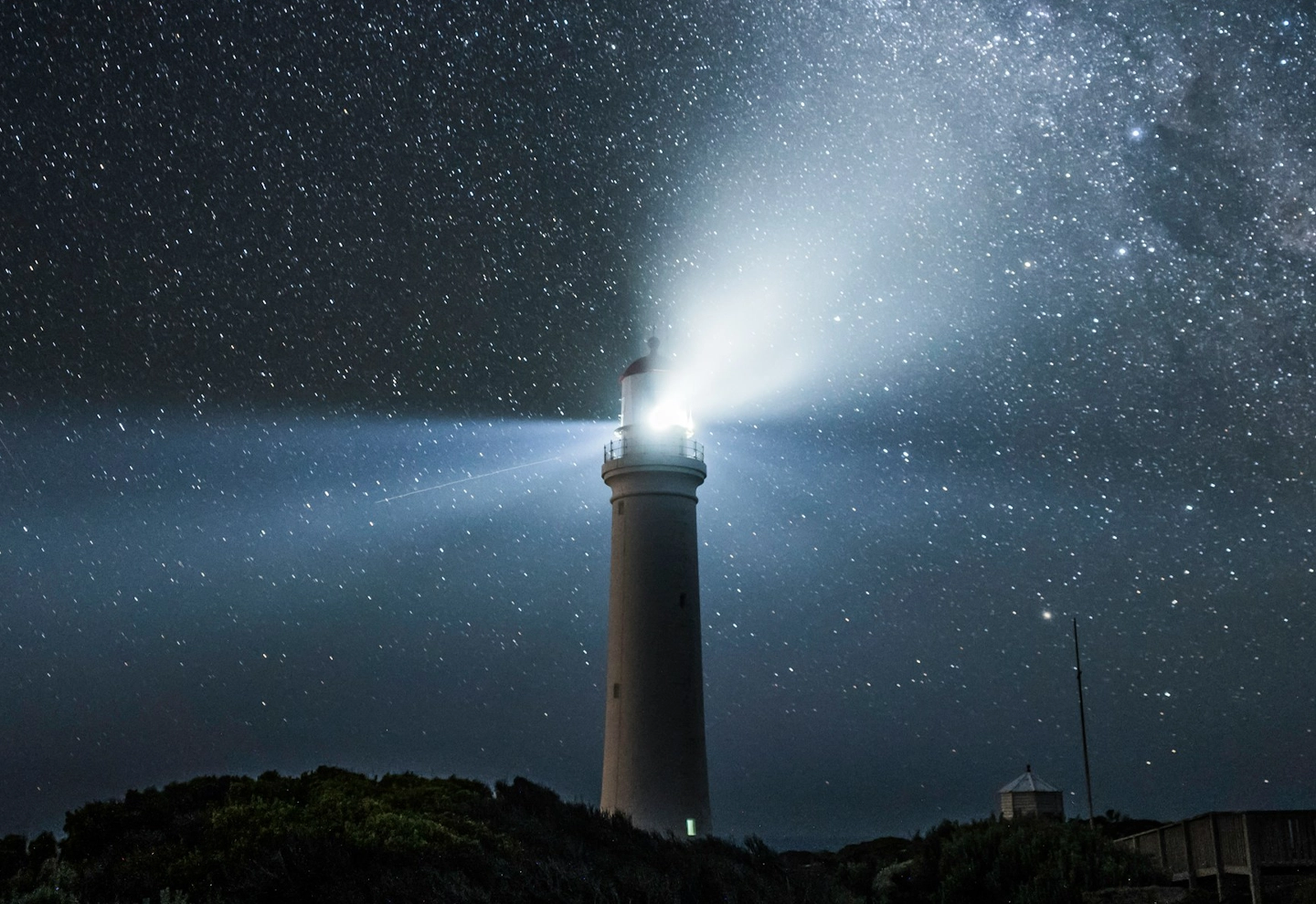 Paisaje de noche estrellada iluminada por la luz de un faro sobre una colina.