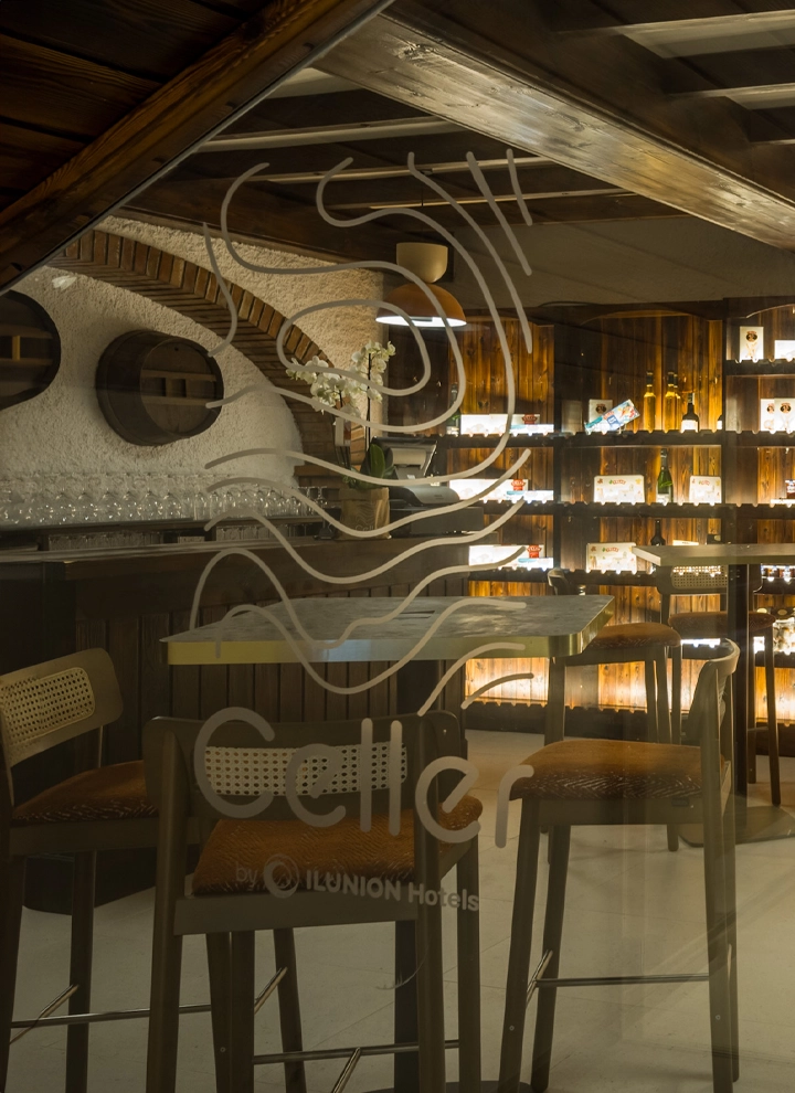 Sala de una bodega con mesas altas y estanterías repletas de botellas de vino colocadas de forma elegante con una luz tenue. sobre el cristal se puede leer Celler by ILUNION Hotels