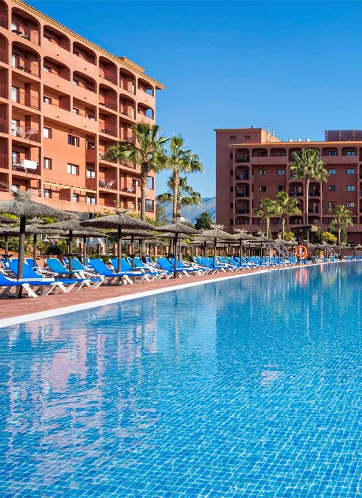Piscina exterior del Hotel ILUNION Miramar, fachada del edificio cuidada de ladrillo marrón, Hamacas a pie de piscina de color azul. Agua cristalina que refleja un cielo azul despejado.