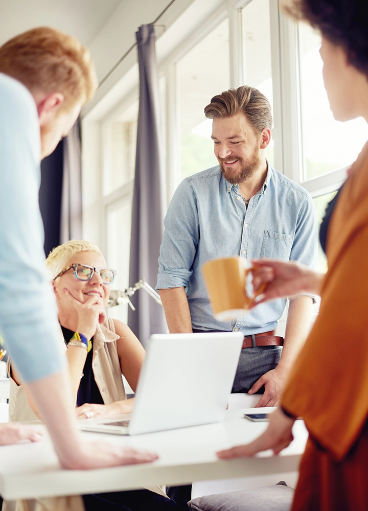 equipo creativo de trabajo, chica con ordenador y chico con barba sonrie frente a otros compañeros