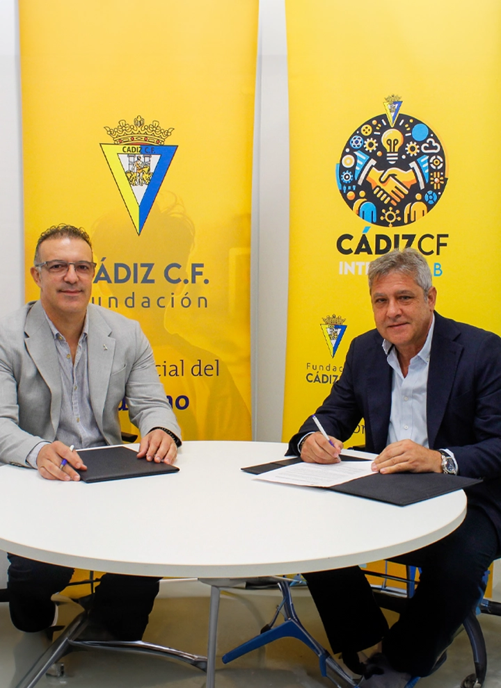 Dos hombres sentados firmando documentos frente a carteles amarillos con logotipos de la Fundación Cádiz CF y el programa de inclusión.