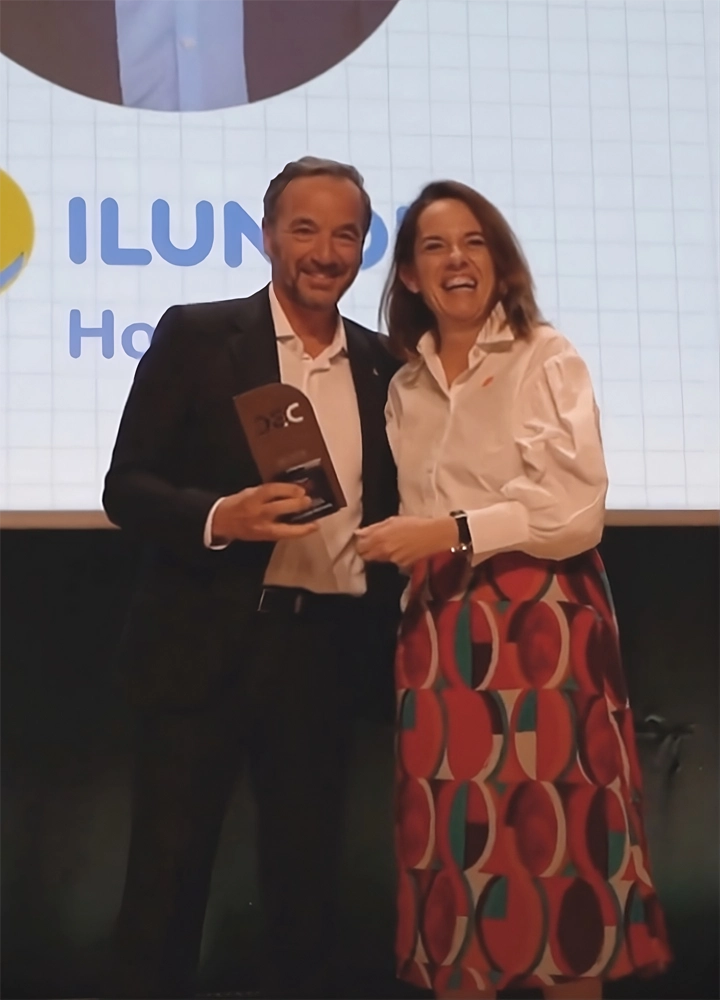 Dos personas de pie en un escenario con una gran pantalla de presentación en el fondo. Una persona lleva un traje y sostiene premio, mientras que la otra viste una camisa blanca y una falda colorida. La pantalla muestra parcialmente el logo de ILUNION Hotels