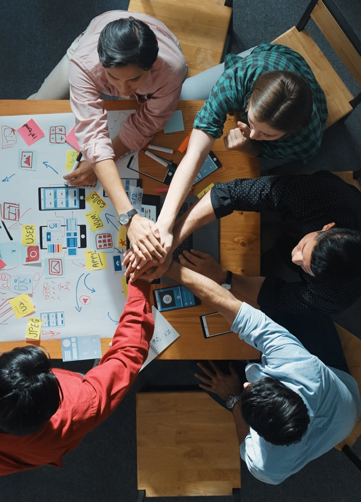 Vista desde arriba de un grupo de personas alrededor de una mesa, con sus manos apiladas en el centro, sugiriendo trabajo en equipo o unidad. La mesa está cubierta con varios papeles que parecen tener diagramas y notas, lo que podría indicar una sesión de planificación o lluvia de ideas.