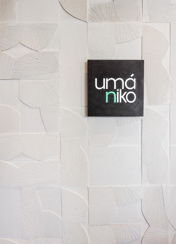 Un panel decorativo de fondo con un diseño geométrico en tonos blancos y gris claro, compuesto por formas semicirculares y líneas texturizadas que crean un patrón abstracto. En el centro, hay un letrero cuadrado oscuro con el texto 'umániko', en el que las letras 'umá' están en blanco y la letra 'n' en un tono verde brillante, seguida de 'iko' en blanco.