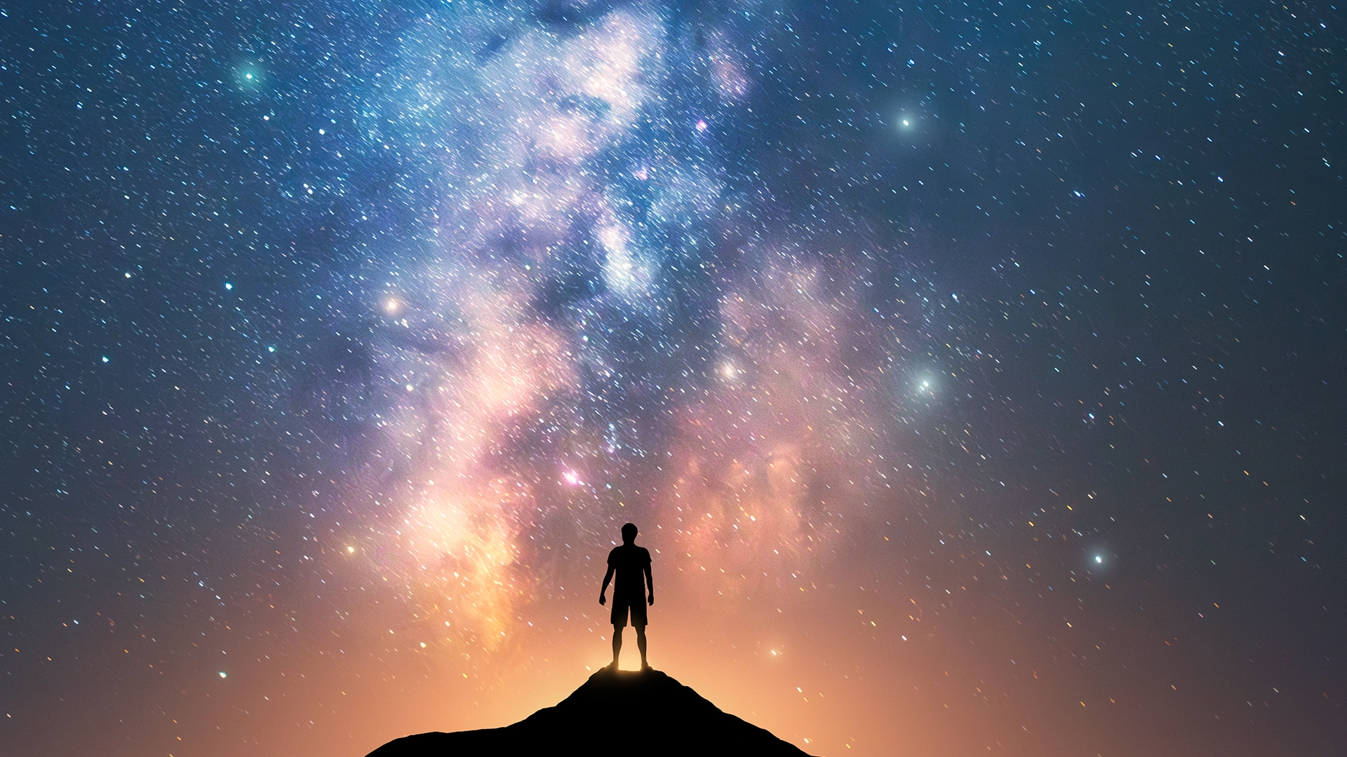 Imagen de la silueta de una persona de pie en la cima de una colina contra un cielo nocturno lleno de estrellas y la Vía Láctea. La vastedad de la galaxia y el pequeño tamaño de la figura humana presentan un contraste interesante, destacando la enormidad y belleza del universo en comparación con un individuo.