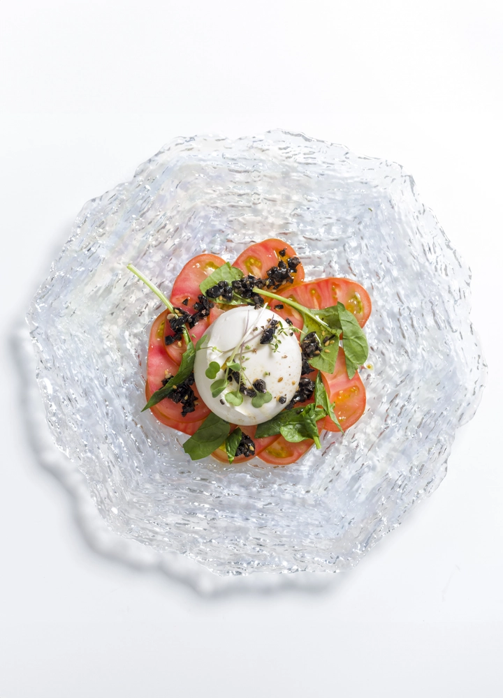 Un plato gourmet presentado en un elegante plato de vidrio con bordes texturizados. En el centro, una burrata fresca se acompaña de rodajas de tomate maduro, hojas verdes y un toque de caviar negro, todo adornado con microvegetales para un acabado sofisticado y colorido.