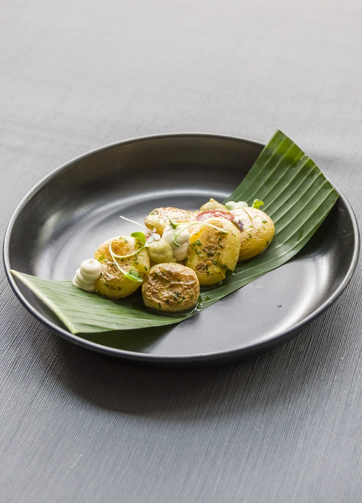 La imagen presenta un plato de papas asadas, servido de manera creativa y visualmente atractiva sobre una hoja de plátano. La iluminación y la composición de la imagen resaltan los colores y las texturas de los alimentos.
