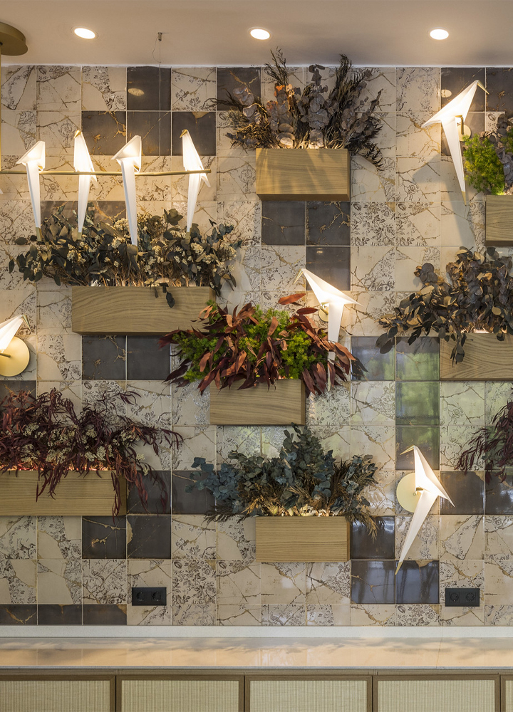 Pared interior contemporánea del restaurante Umaniko Atrium, con un diseño modular que combina baldosas de cerámica con tonalidades neutras, macetas de madera con diversas plantas y lámparas de pared en forma de pájaro de papel.